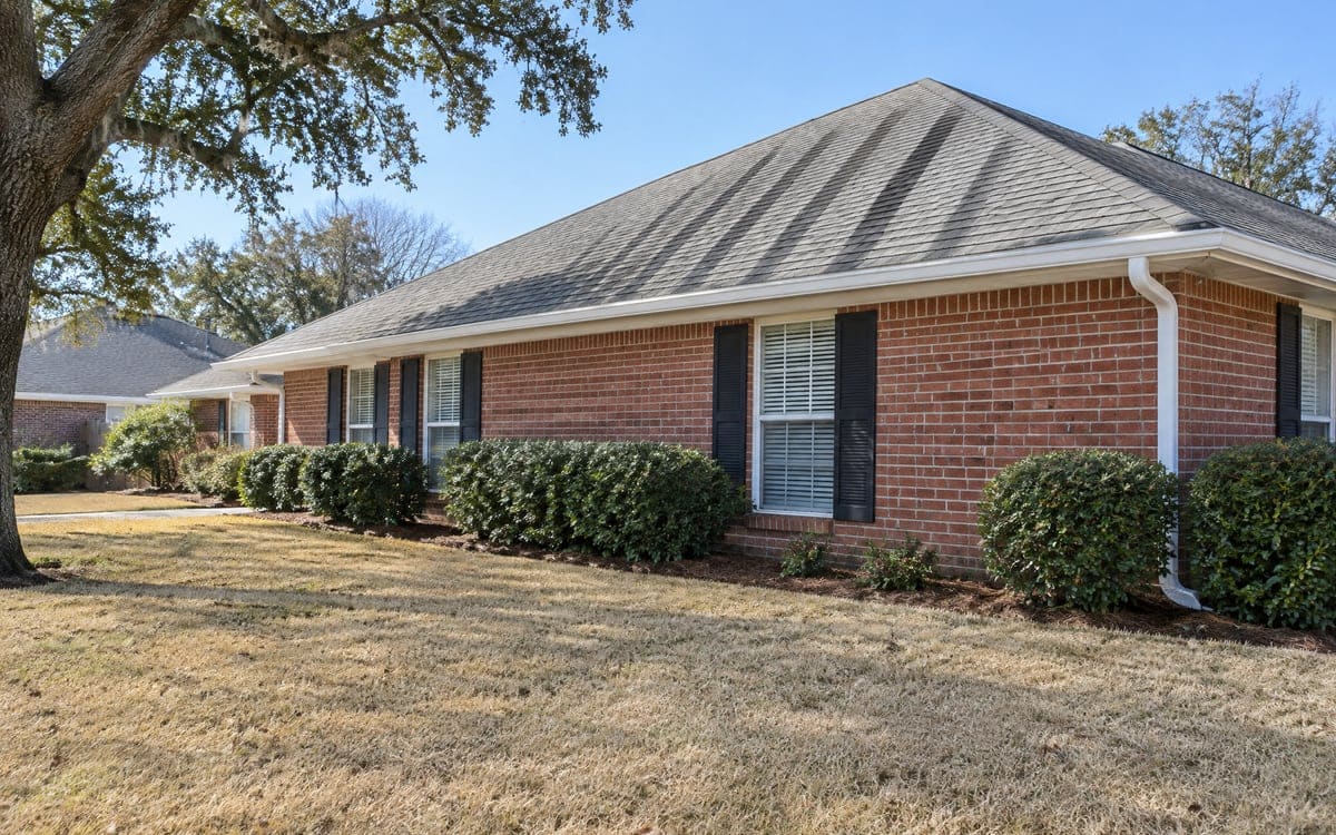 A brick ranch's hip roof seen from the yard on a winter day, with long dark bands running down the gray shingles from the ridge to the gutter and the lawn tan behind the shrubs.