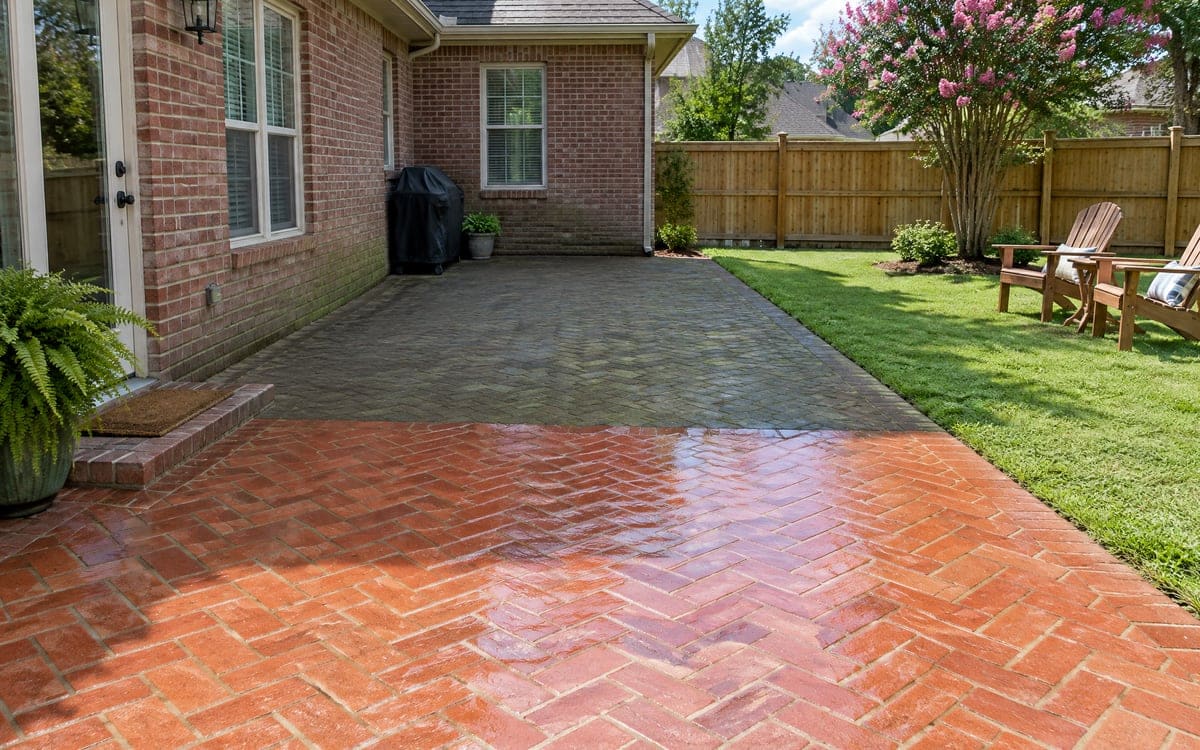 A near red-brick herringbone patio sits bright after a wash, with a gray concrete-paver patio behind it still shade-dulled, a lawn and wood fence beyond and Adirondack chairs on the grass.