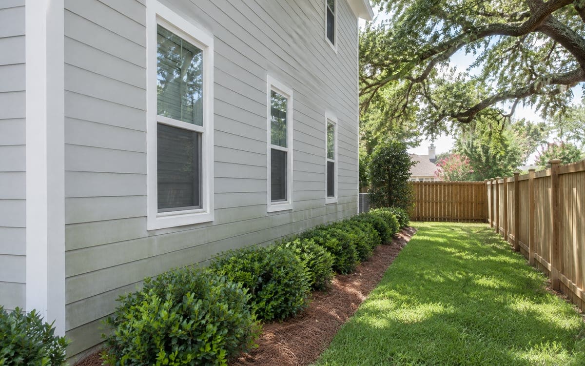 Pale gray lap siding runs along the shaded side of a two-story house, with a faint gray-green film on the lower boards above a bed of trimmed boxwoods and a wood fence.