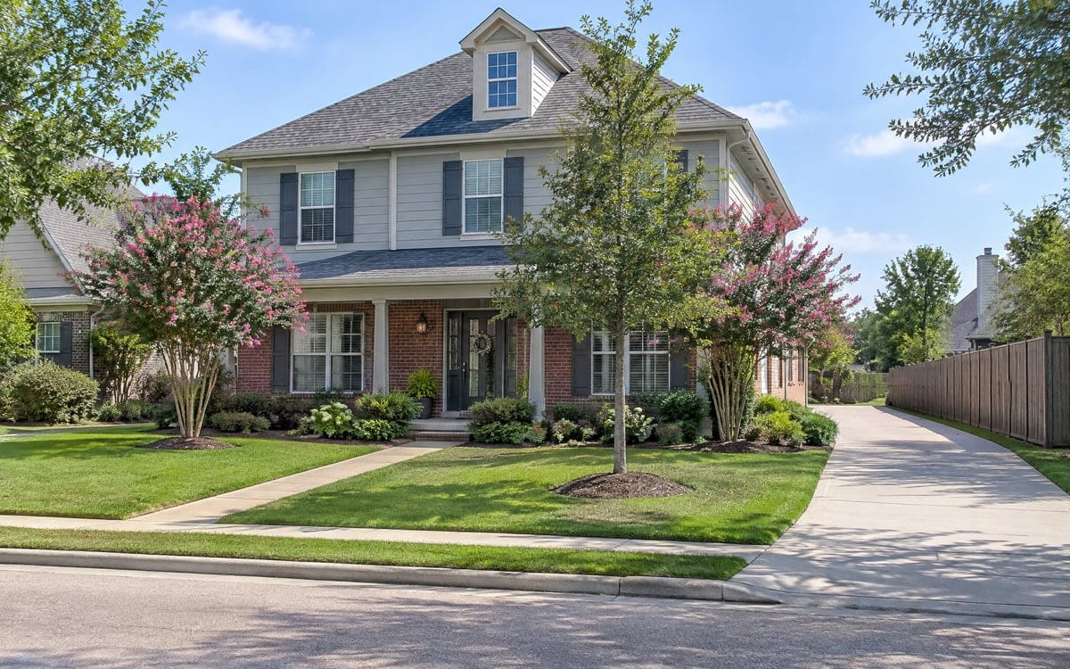 A two-story house with gray lap siding over a brick base, a mown front lawn and a long concrete driveway down one side, the kind of home a pressure washing cost gets quoted for.