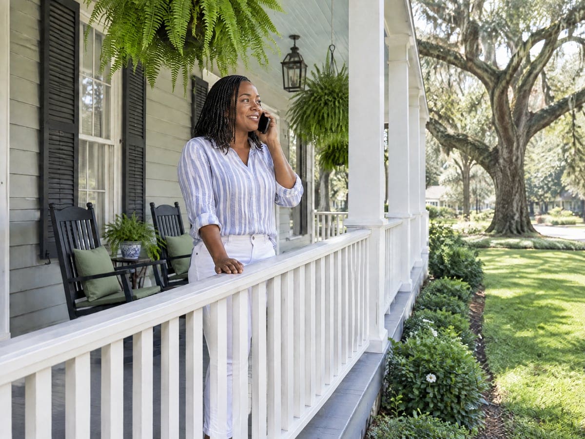 A woman on a long wraparound porch takes a call on her phone, framed by hanging ferns, white spindle railing and live oak branches over the lawn beyond.