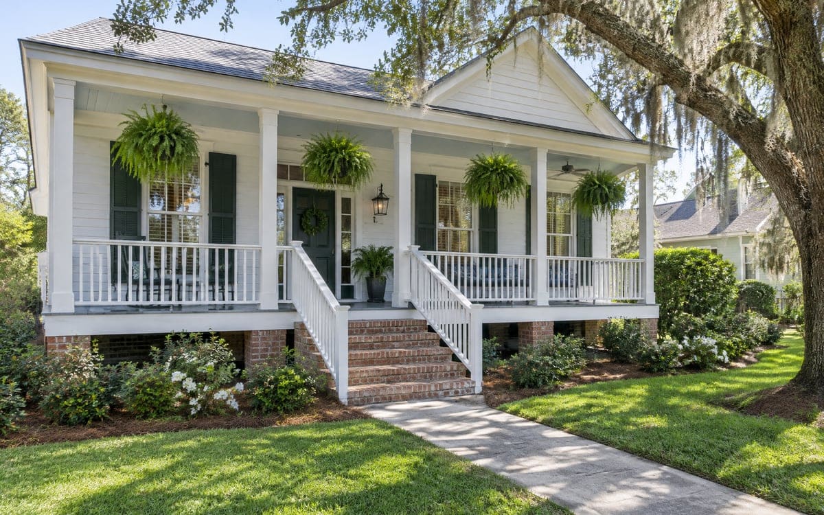 A white raised cottage with a deep front porch, four hanging ferns and dark green shutters sits under a live oak draped with Spanish moss, azaleas and a brick walk at the front steps.
